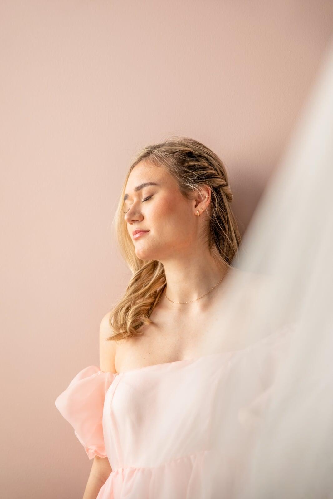 Soft portrait of a blonde model in a pale pink off-shoulder tulle dress, posing with eyes closed against a blush pink background with sheer fabric in the foreground.