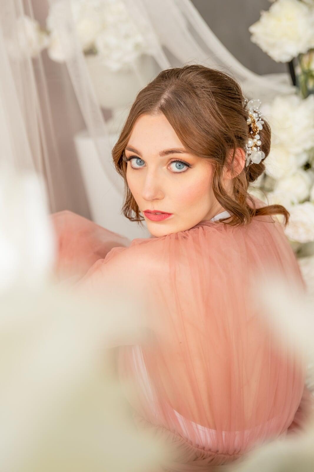 Soft bridal portrait of a model in a blush pink tulle dress, looking over her shoulder with romantic updo styling, pearl floral hair accessories, and soft glam makeup, surrounded by white flowers and delicate draping.
