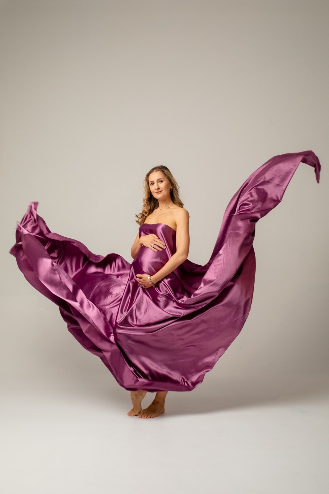 Pregnant woman standing in a studio wrapped in flowing purple satin fabric, holding her baby bump as the fabric moves around her dramatically.