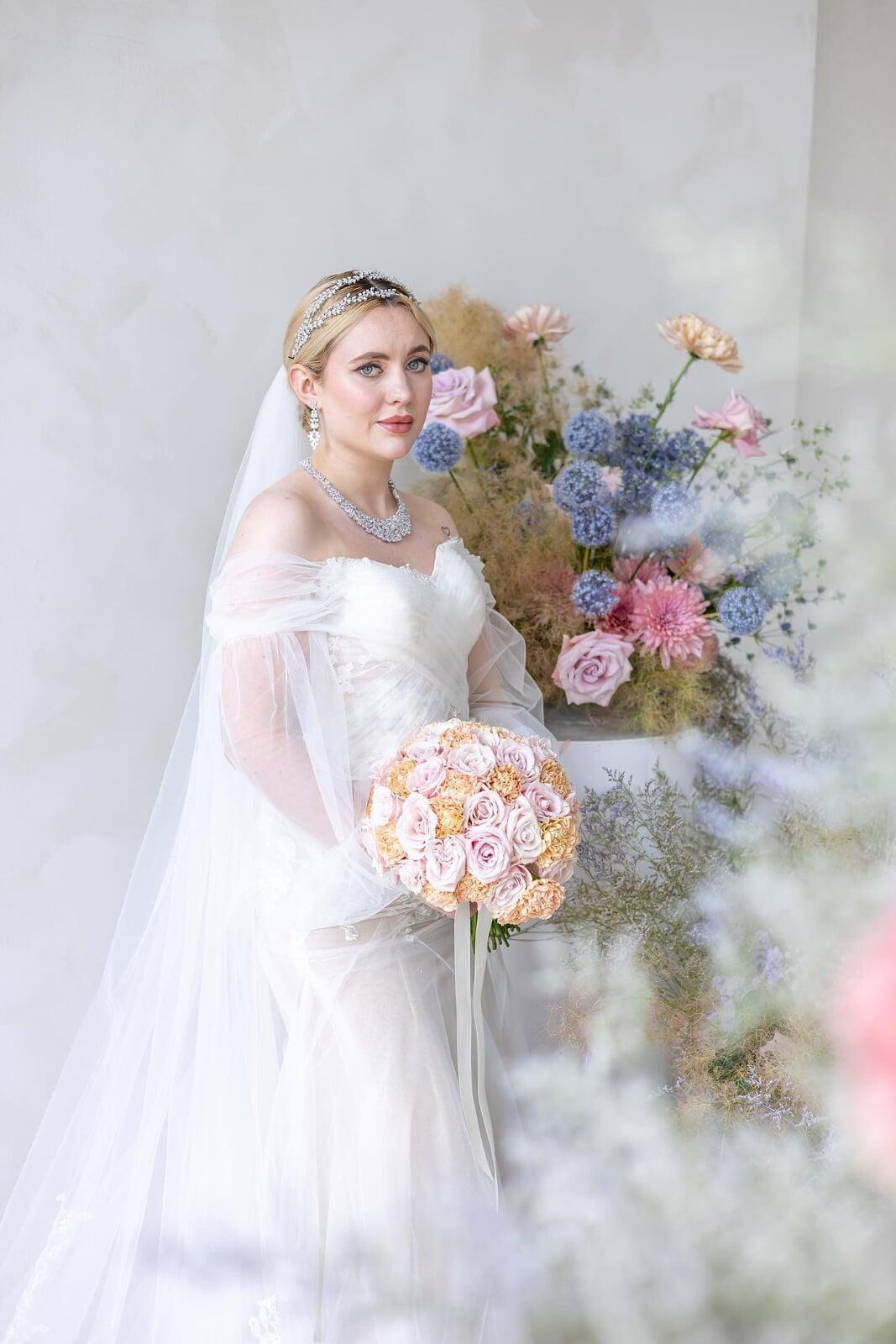 Bride in soft glam bridal makeup wearing a white wedding gown and veil, holding a pastel bouquet beside romantic floral arrangements.