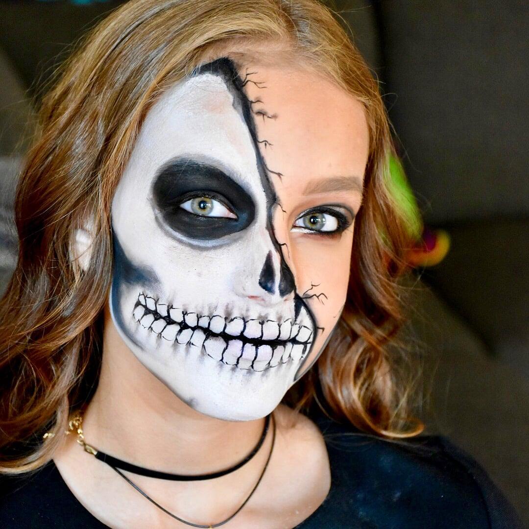 Close-up portrait of a model with half-face skull makeup, featuring white and black skeletal details, cracked face paint, smoky eye makeup, and wavy hair against a dark background.