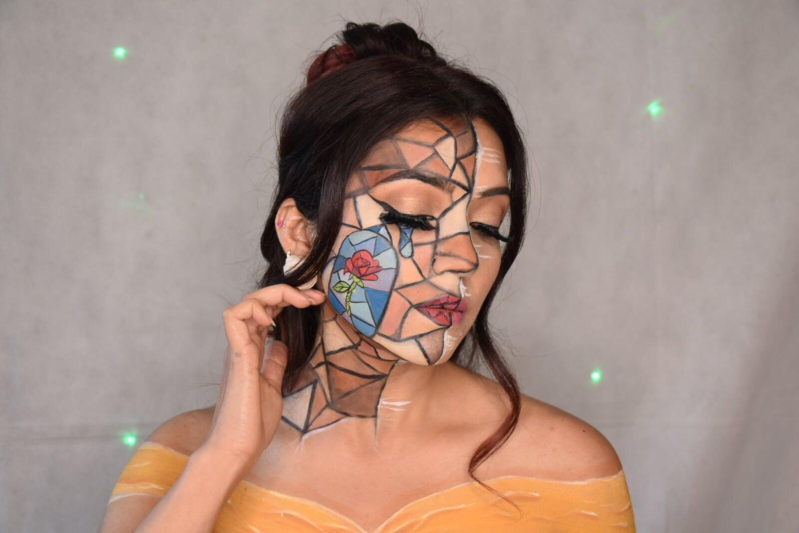 Portrait of a model with creative stained-glass inspired face and body makeup, featuring cracked geometric lines, a painted blue rose on the cheek, soft glam eye makeup, and an off-shoulder yellow design against a grey background.