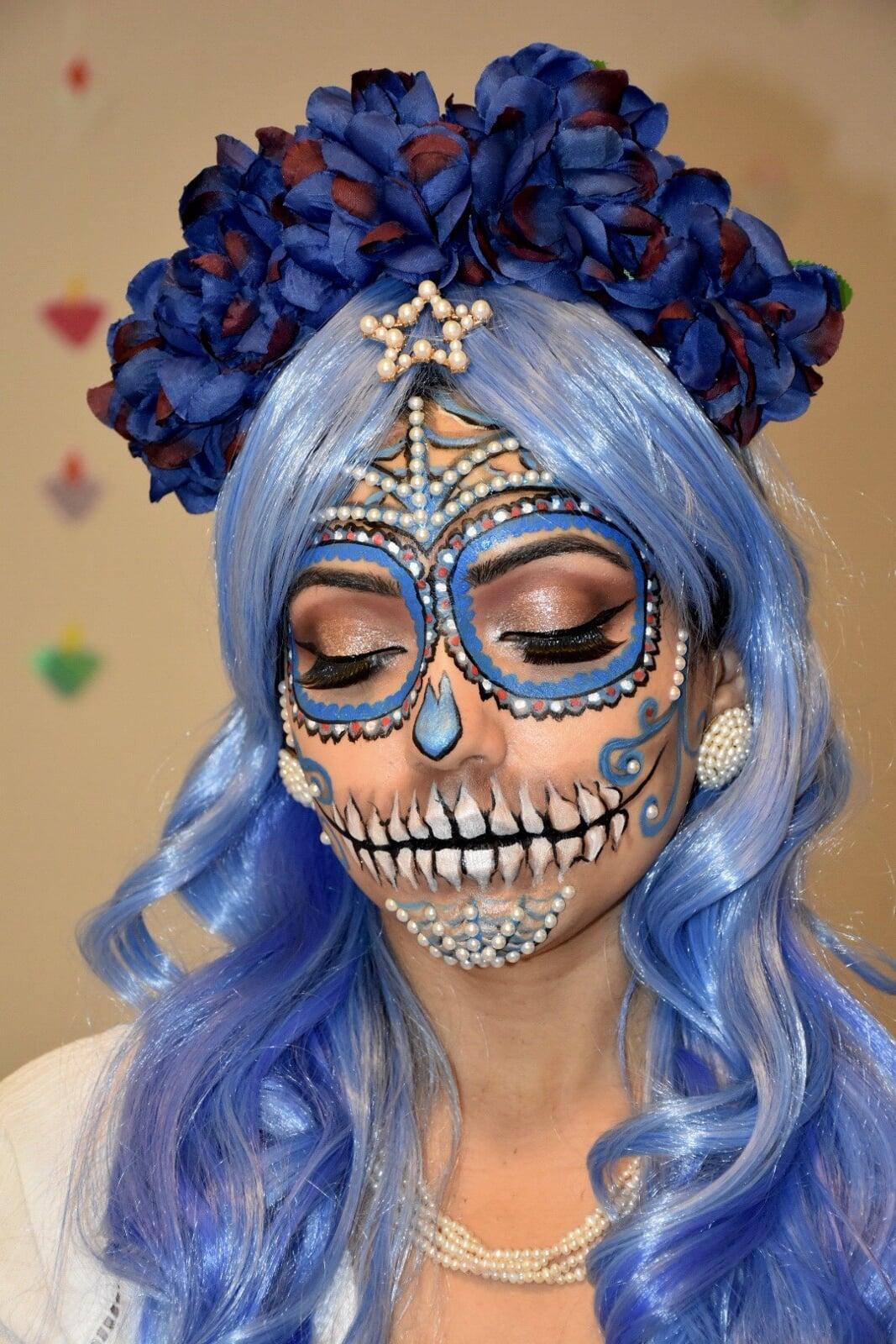Portrait of a model with blue sugar skull makeup, pearl and rhinestone accents, closed eyes, blue wavy hair, and a floral headpiece with dark blue flowers.