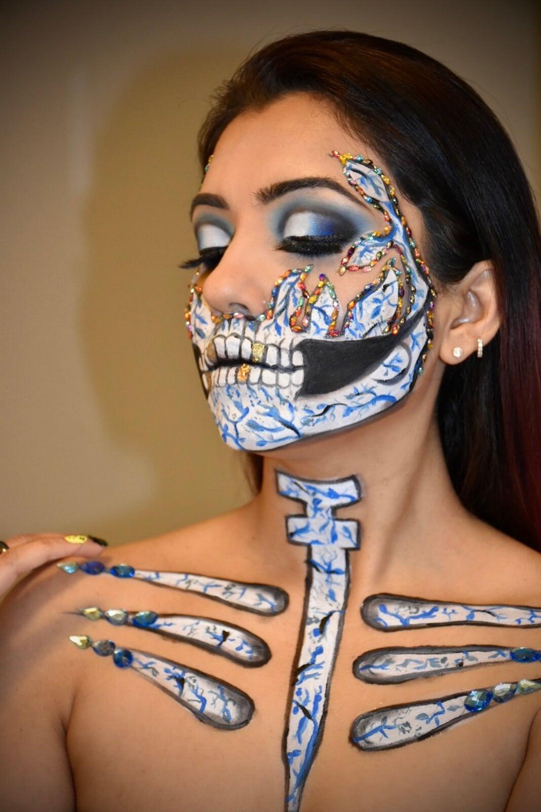 Portrait of a model with elaborate blue-and-white sugar skull body art, featuring skeletal face, neck, and collarbone makeup with floral details, rhinestone accents, dramatic blue eyeshadow, and eyes closed.