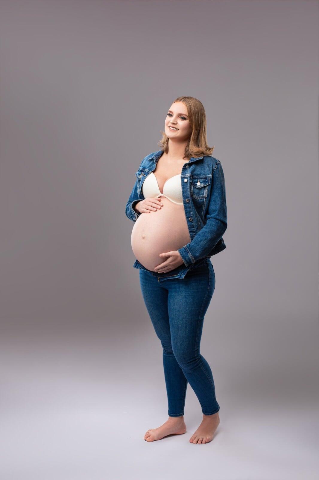 Pregnant woman standing in a studio wearing jeans, a denim jacket, and a white bralette, gently holding her baby bump against a grey background.