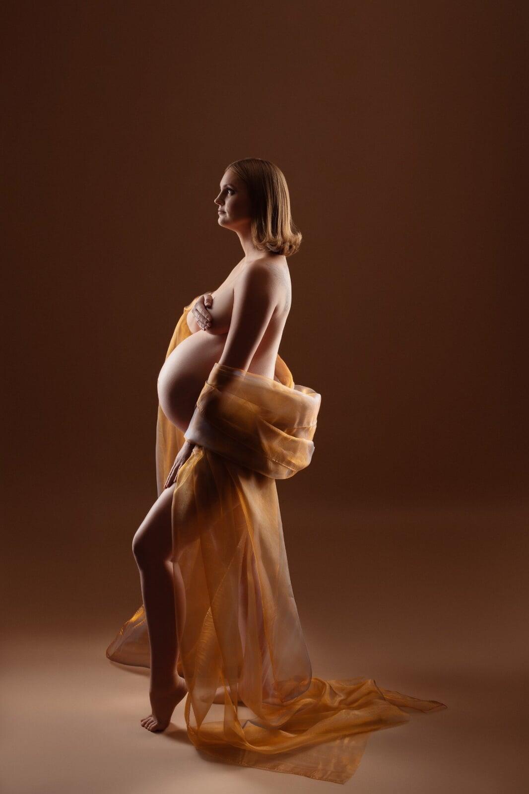 Side-profile maternity portrait of a pregnant woman wrapped in sheer golden fabric, softly lit against a warm brown studio background.