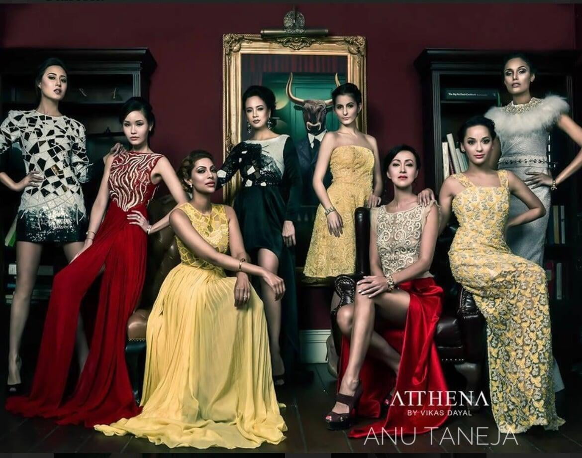 Editorial group fashion portrait of seven models in elegant evening gowns and embellished outfits, posed in a dark, luxurious room with red walls, bookshelves, leather chairs, and framed artwork in the background.