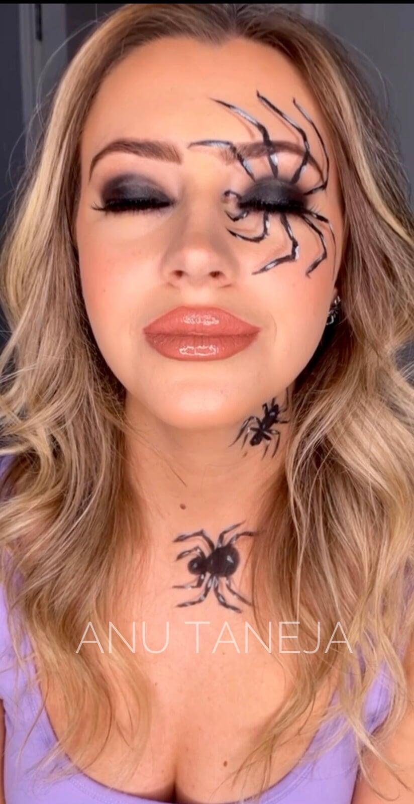Close-up portrait of a model with blonde waves, glossy nude lips, smoky eye makeup, and painted black spider designs around one eye, on the neck, and on the chest.