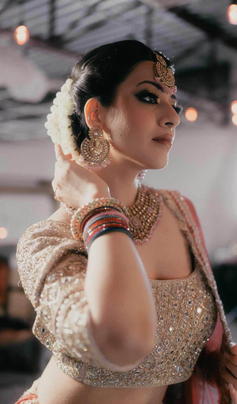 Side-profile bridal portrait of a woman wearing South Asian bridal jewelry, soft glam makeup, floral hair adornment, and an embellished gold blouse, gently touching her hair against a softly lit indoor background.