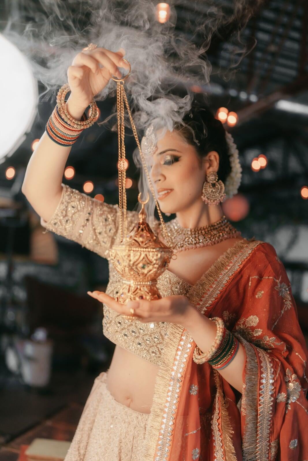 South Asian bridal portrait of a woman in an embellished lehenga and red dupatta, holding a smoking gold incense lantern while wearing traditional jewelry, bangles, and soft glam makeup in a warmly lit indoor setting.