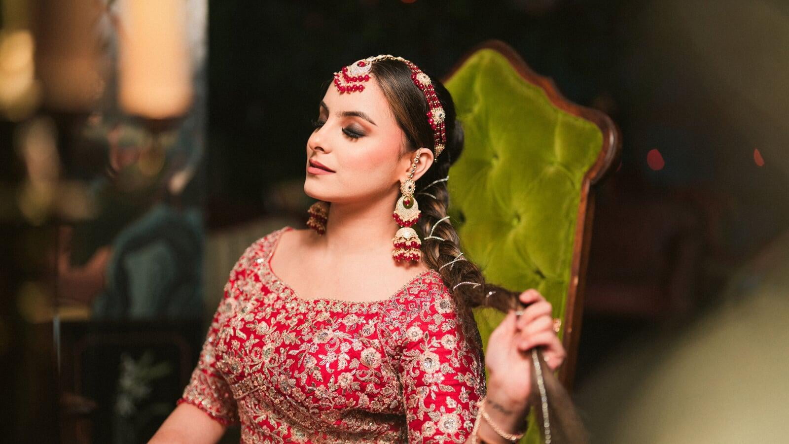 Full-length outdoor bridal portrait of a woman wearing a heavily embellished blush pink lehenga with a matching dupatta and statement jewelry, standing beneath an ornate stone archway.